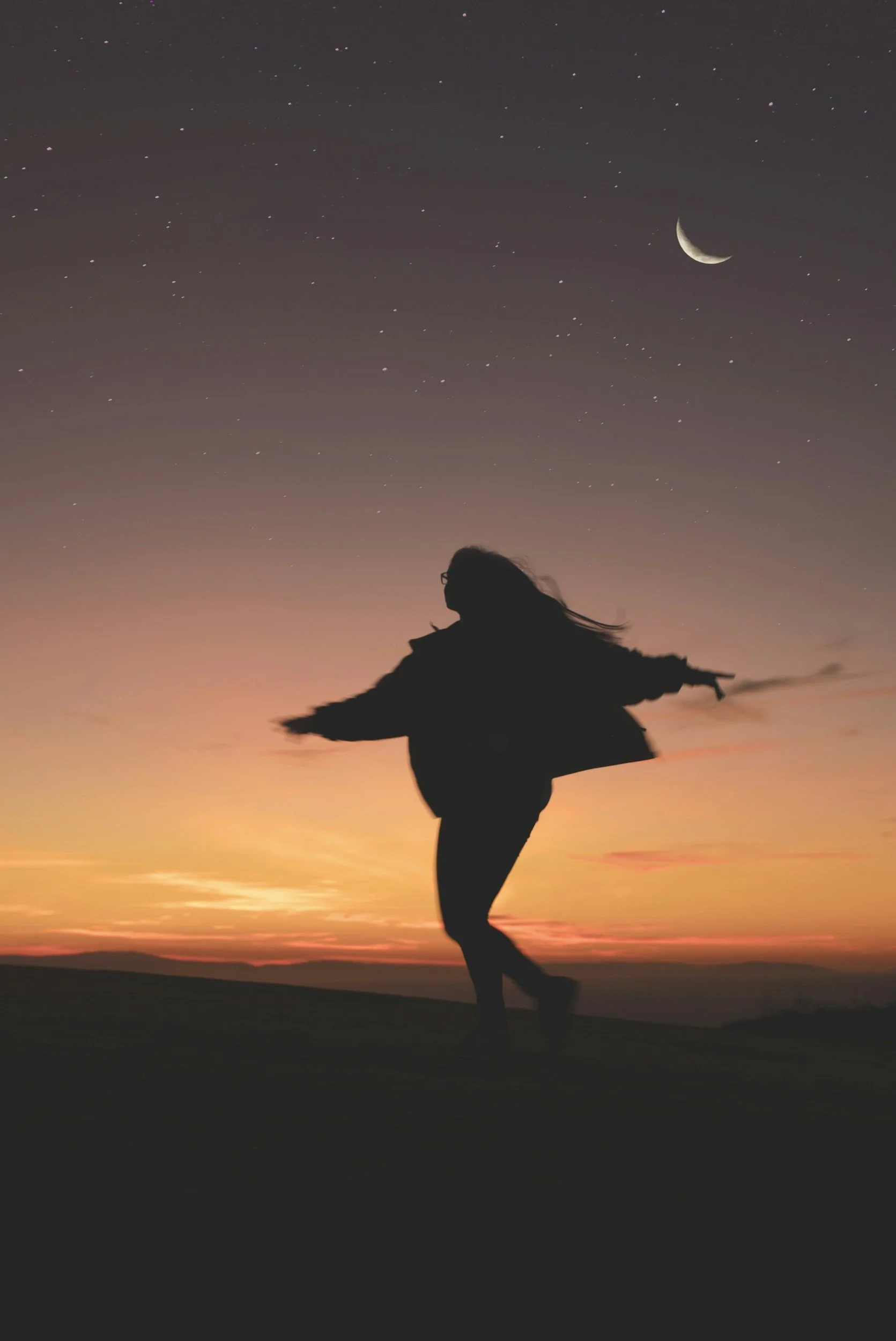 Silhouette of a woman dancing freely under a dusk sky with a crescent moon, arms open — an image of what it feels like to raise your vibrations and let your radiance move through you.
