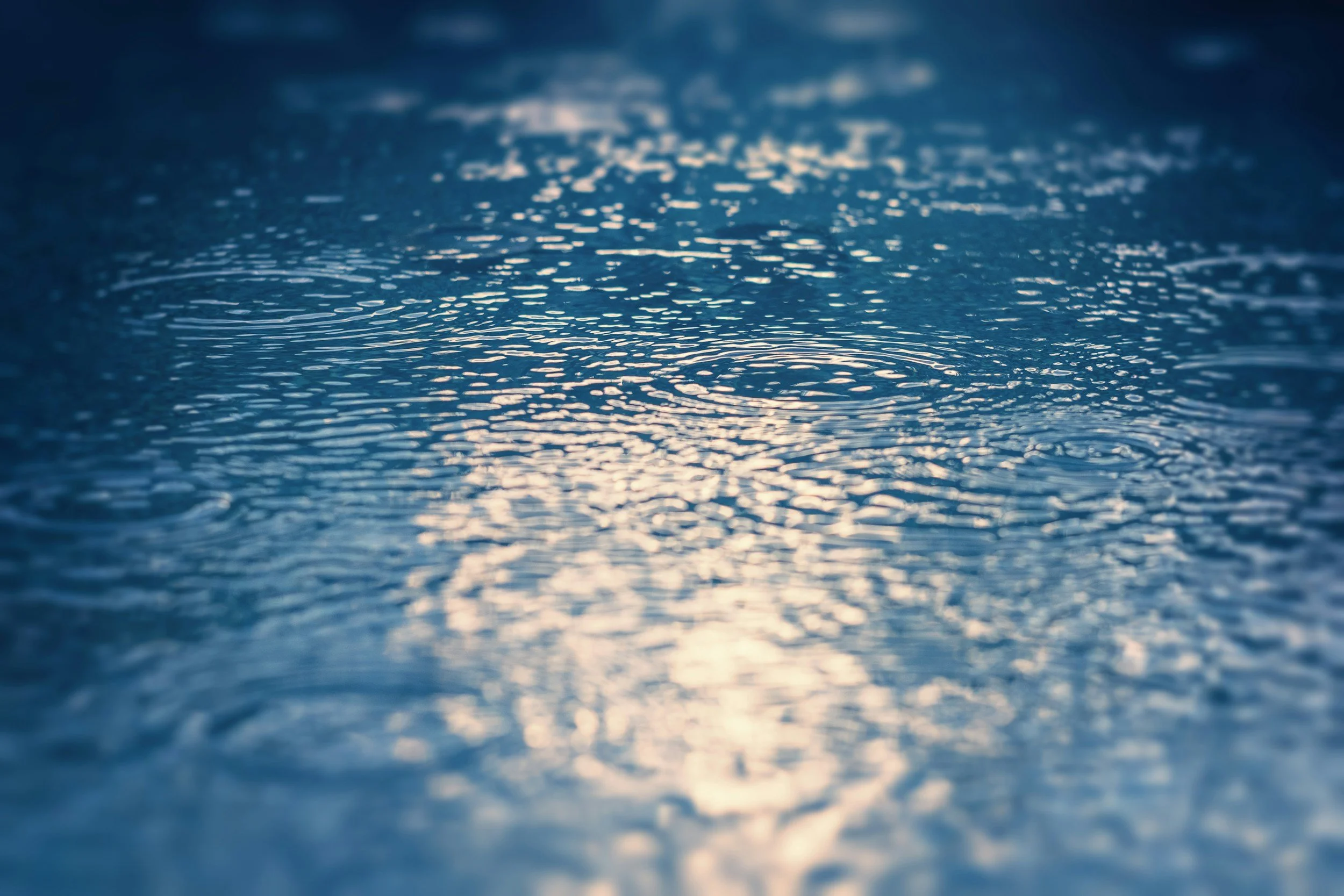 Raindrops rippling across a dark, glassy water surface with soft light reflecting through — an image of healing perfectionism, of what clears when you stop trying to control the surface.