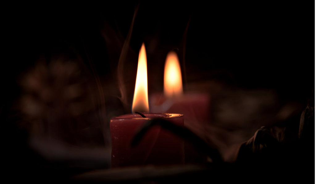 Two dark red candles burning steadily in near-total darkness, their flames the only light against the black: an image of shadow work and healing perfectionism, staying with what's dark rather than looking away from it.