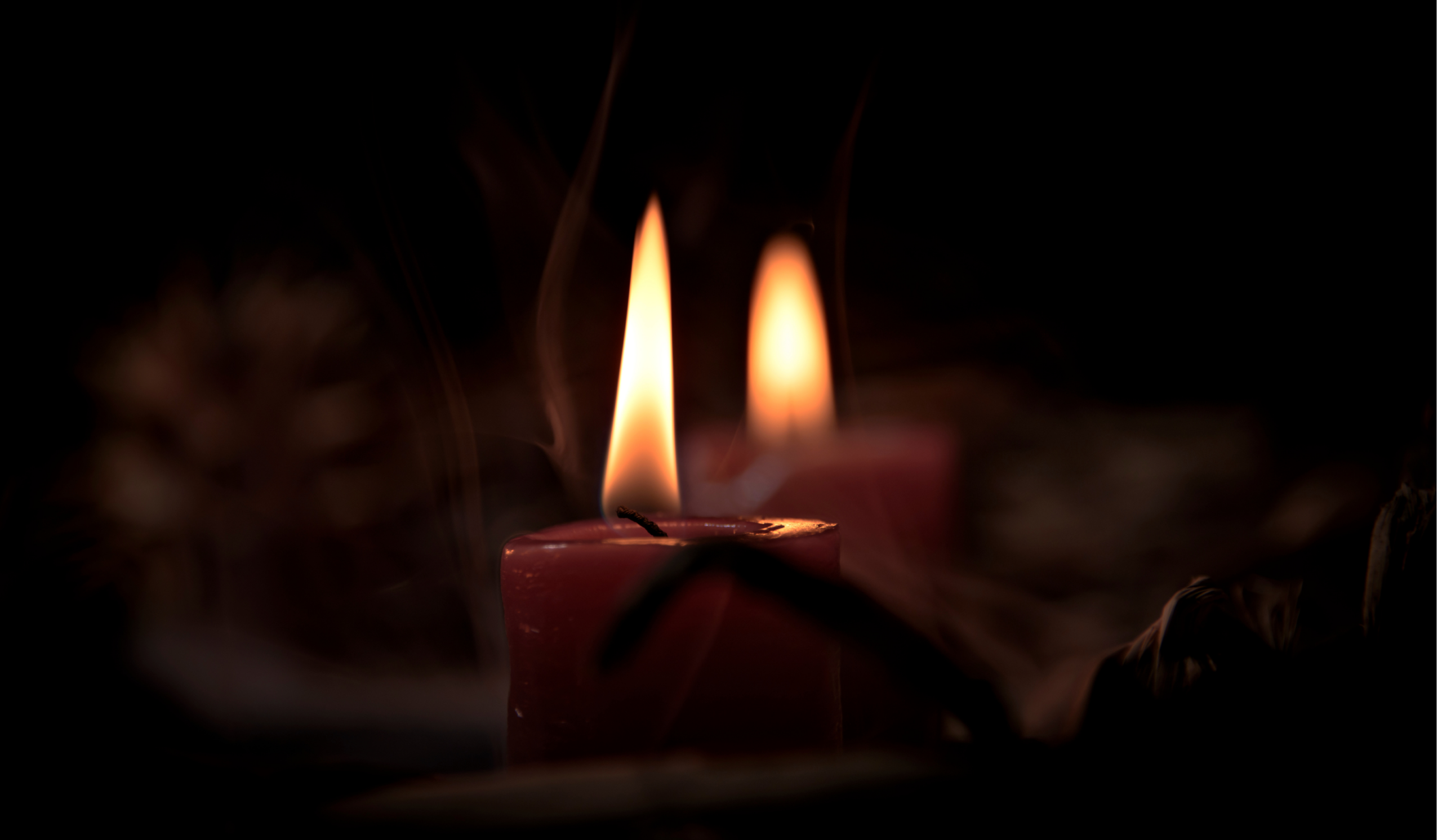 Two dark red candles burning steadily in near-total darkness, their flames the only light against the black: an image of shadow work and healing perfectionism, staying with what's dark rather than looking away from it.