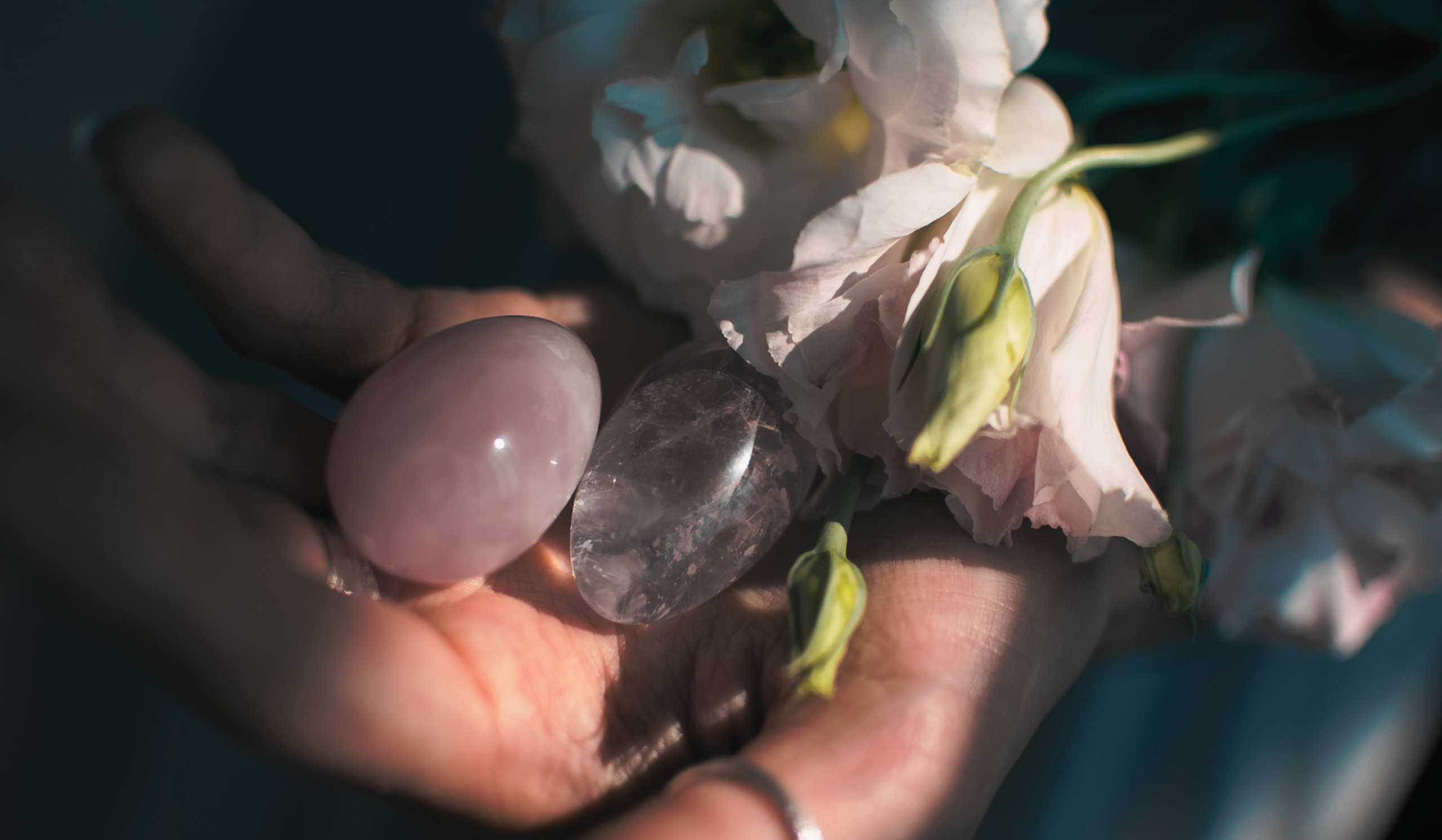 Rose quartz yoni egg and smoky quartz stone cradled in an open palm, surrounded by soft white blooms.