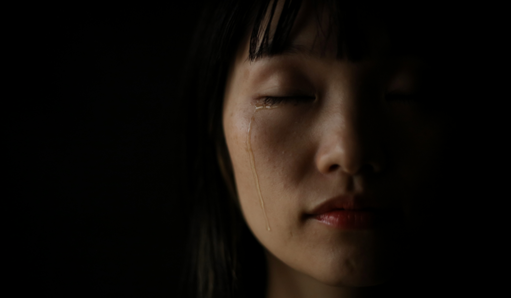 Woman with eyes closed, a single tear falling in the dark, the quiet exhaustion behind people pleasing and good girl conditioning.